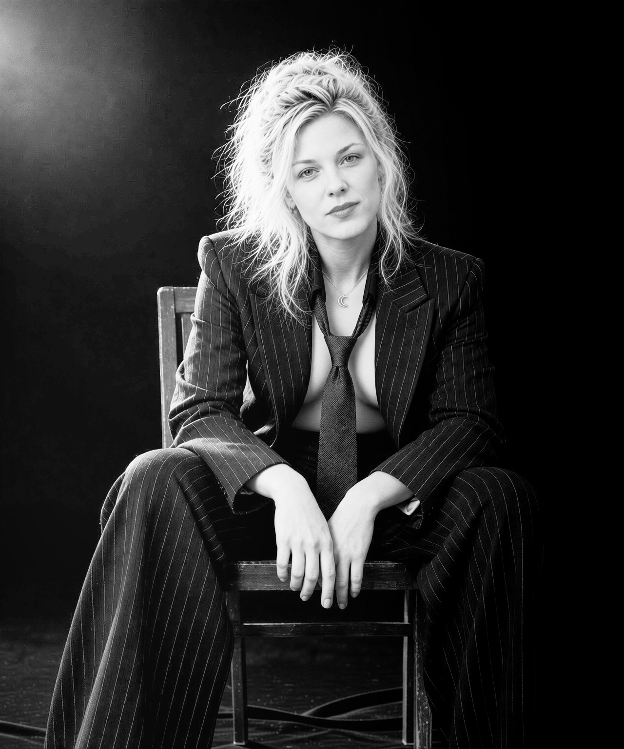 Black-and-white seated portrait of Clarapetra leaning forward on a wooden chair against black, in a pinstripe suit and loose tie.