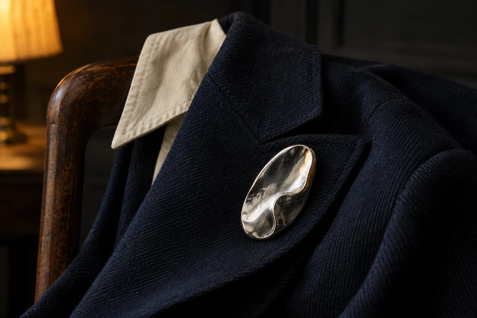 AI editorial concept: one sculptural silver-colored brooch rests on the lapel of an ink-blue wool jacket with an ivory collar in warm lamplight.
